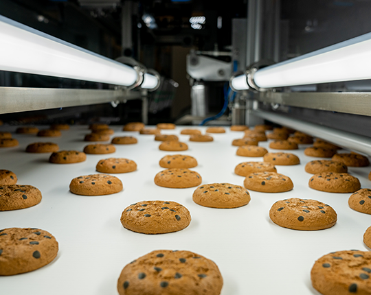 Chocolate chip cookies moving through a Syntegon robotic assisted conveyor system for final packaging.